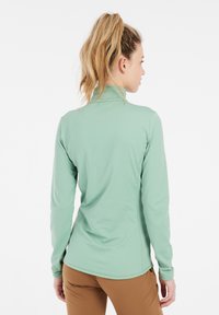 Maglietta sportiva a maniche lunghe di colore verde menta con colletto alto, caratterizzata da una superficie liscia e un design aderente, abbinata a pantaloni marroni aderenti.