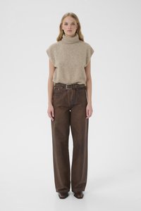 Frau trägt beige ärmellosen Rollkragenpullover und hoch taillierte, lockere braune Hose mit Gürtel, steht vor einfarbigem Hintergrund.