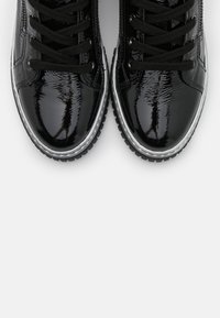 Un par de zapatos de cuero negro brillante con cordones negros y suelas gruesas de taco blanco y negro sobre un fondo blanco.