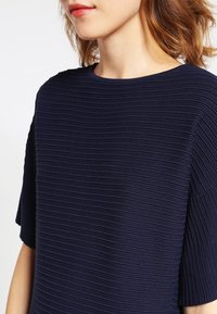 Nainen, joka käyttää tummansinistä ribattua lyhythihaista toppia, jossa on pyöreä kaula-aukko, näkyen osittain hänen kasvonsa ja olkapitkät hiukset.