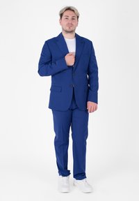 Abito blu su misura con rever a profilo, due bottoni e tasche laterali. Abbinato a sneakers bianche e una t-shirt bianca sotto.