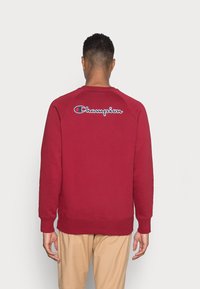 Felpa maroon con maniche raglan, polsini e vita a coste; presenta il logo "Champion" ricamato sul retro. Tessuto morbido e testurizzato.