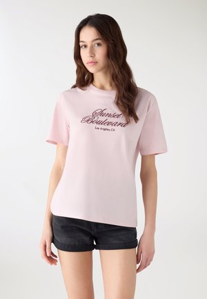 Giovane donna con capelli castani lunghi che indossa una maglietta rosa chiaro con la scritta "Sunset Boulevard Los Angeles, CA" e pantaloncini di jeans neri.
