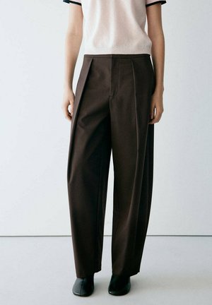 Persona que lleva pantalones marrón oscuro de tiro alto y pernera ancha, camiseta de punto beige claro de manga corta y zapatos negros, de pie frente a un fondo blanco liso.