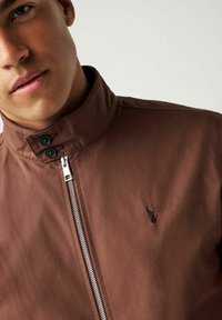 Chaqueta ligera marrón con cuello alto, cierre lateral y logo bordado en el pecho. Textura suave con detalles de botones.