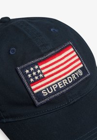 Gorra de béisbol azul marino con un parche rectangular que muestra una bandera estadounidense bordada y el texto "SUPERDRY" debajo. Material de algodón, diseño estructurado.