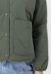 Grøn quiltet jakke med bølgende syninger, trykknapper og en frontlomme. Stoffet ser glat ud; ærmerne er let samlet ved manchetterne.