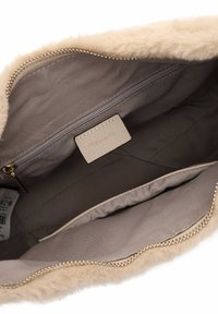 Intérieur de sac à main beige clair avec une texture douce semblable à de la fourrure, comportant une poche zippée, une doublure lisse et une petite étiquette de marque. Fermeture éclair en ton doré.
