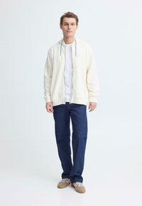 Sweat à capuche crème clair zippé avec cordons bleus, superposé sur un t-shirt blanc. Associé à un jean droit bleu marine et des baskets beiges.