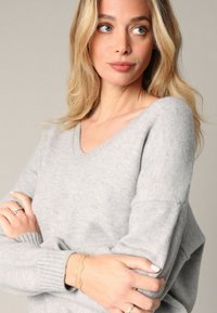 Pull léger gris à col en V avec des manches longues, présentant une texture douce et des poignets côtelés. Les mains du modèle sont ornées de bagues et d'un bracelet.