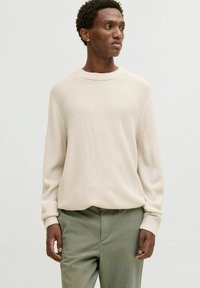 Light beige knit sweater with a round neckline and ribbed cuffs, paired with olive green pants. Textured fabric and relaxed fit.