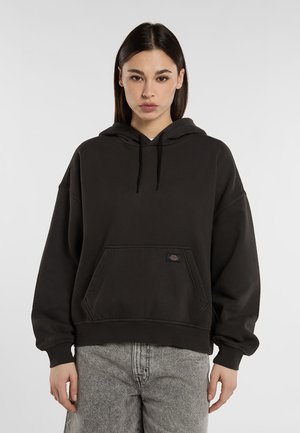 Ung kvinna med rakt mörkt hår bär svart hoodie och ljusgrå jeans, stående mot en ljus bakgrund.