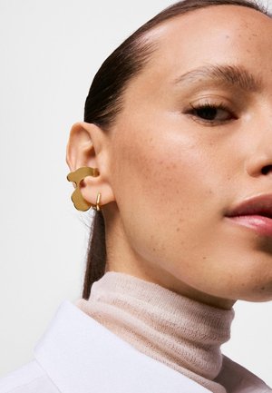 Nahaufnahme eines Frauengesichts, das ihr Ohr mit einem goldfarbenen abstrakten Ohrstulpen und kleinem goldenen Creolenohrring zeigt, sie trägt einen leichten Rollkragenpullover und ein weißes Hemd.