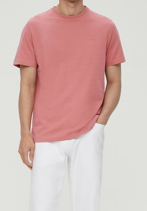 Mann trägt ein schlichtes staubig rosa kurzärmeliges T-Shirt und weiße Hose, steht mit einer Hand in der Tasche vor neutralem Hintergrund.