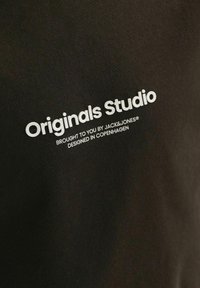 Juodas audinio marškinėliai su ryškiai balta užrašu: "Originals Studio" ir mažesniu tekstu žemiau, nurodančiu "pateikė JACK&JONES®."