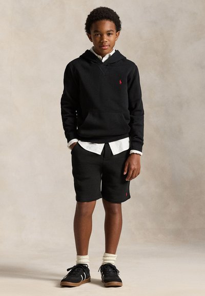 Chlapec stojaci čelom vpredu, oblečený v čiernej kapucni, bielom tričku, čiernych šortkách, bielych ponožkách a čiernych teniskách na jednolícom pozadí.