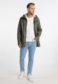 Olivenfarvet vandtæt jakke med grå hætte, trykknapper og sidelommer, båret over en hvid T-shirt og lyseblå jeans.