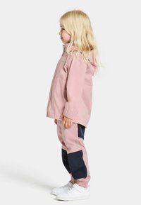 Ljusrosa vattentät jacka med huva, ihop med matchande rosa och marinblå byxor. Har elastiska ärmslut och vita sneakers.