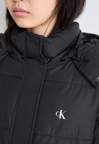 Melnā pūkainā jaka ar augstu apkakli, ar vertikālām quiltēšanām un baltu "CK" logo kreisajā krūtī. Gluda tekstūra.