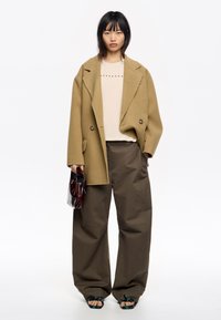 Abrigo de lana marrón de corte oversized y doble botonadura sobre una blusa color crema con tachuelas decorativas, combinado con pantalones khaki anchos y sandalias gruesas.