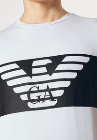 Person wearing a white T-shirt with a large black stylized eagle logo and "GA" letters printed across the chest.