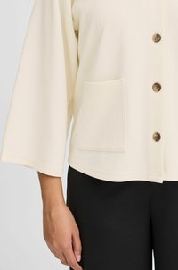 Blusa texturizada color blanco roto con mangas tres cuartos, cierre de botones y un bolsillo frontal. Combinada con pantalones negros.