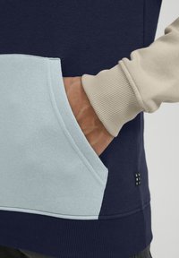 Suéter en azul marino con un bolsillo frontal de color azul claro. Presenta un puño y dobladillo de canalé beige. Textura suave con detalles de costura visibles.
