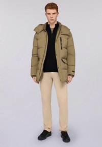 Prošívaná bunda v olivově zelené barvě s kapucí, vybavená zipem a patentními knoflíky, boční kapsy a texturovaný prošitý design. Stylizováno s béžovými kalhotami a černými botami.
