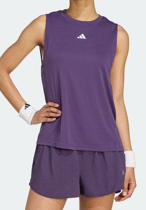 Osoba w bezrękawniku Adidas w kolorze fioletowym, pasujących szortach i białych opaskach na nadgarstki z czarnymi logotypami Adidas, stojąca z jedną ręką na biodrze.