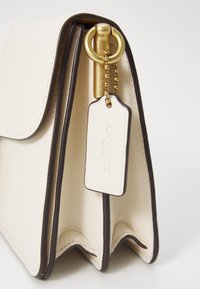 Cream-colored leather handbag with dark brown trim, featuring a gold metal ring and a white tag with "COACH" embossed. Textured finish.
