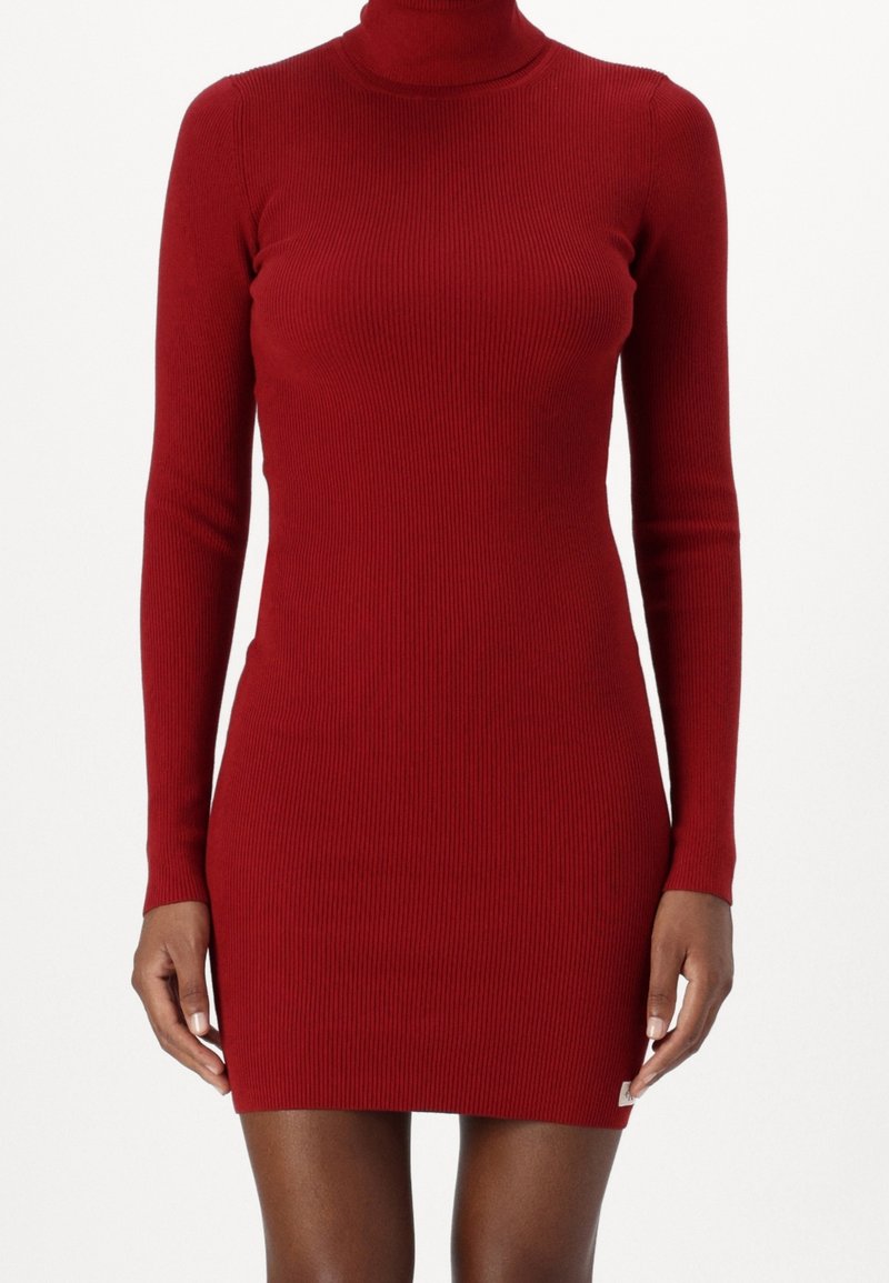 Robe rouge en maille côtelée ajustée à manches longues avec col roulé, portée par une personne à la peau foncée, visible de la mi-cuisse au cou.