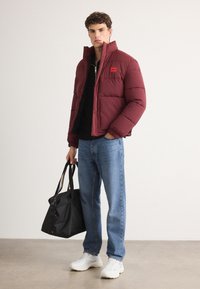 Burgunderfarvet pufferjakke, quiltet design, sort zip-up sweater under, blå jeans, hvide sneakers og en sort taske.