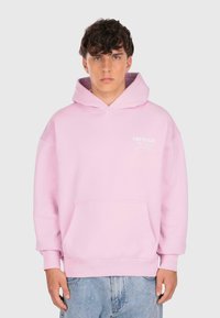 Giovane uomo che indossa una felpa leggera rosa con testo bianco sul petto e jeans blu, in piedi contro uno sfondo grigio chiaro uniforme.