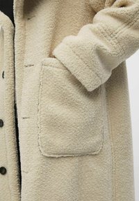 Beige Fleece-Mantel mit strukturiertem Oberflächen, großen aufgesetzten Taschen und Knopfverschluss. Der Stoff hat ein weiches, plüschiges Aussehen.