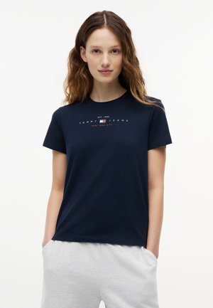 Mujer con cabello castaño ondulado, vistiendo una camiseta azul marino de Tommy Jeans y pantalones gris claro, de pie con las manos en los bolsillos contra un fondo blanco.