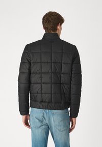 Jachetă puffer neagră matlasată cu guler în picioare, talie elastica și cusături vizibile. Stilizată cu blugi lejeri de culoare albastră deschisă.