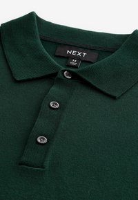 Polo de color verde oscuro con un tejido texturizado, que presenta un cuello, tres botones grises y una etiqueta con la marca "NEXT" y tamaño "M".