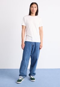Pessoa com cabelo preto longo está vestida com uma t-shirt branca simples, calças de ganga azuis largas e ténis verdes e brancos, em um piso azul.