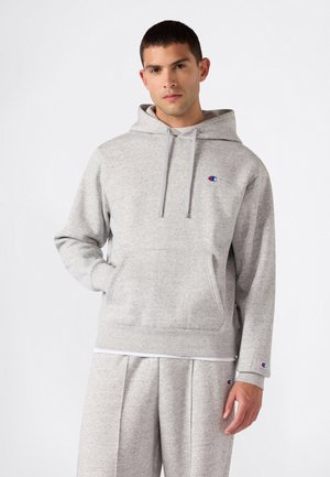 Uomo che indossa una felpa con cappuccio Champion grigio chiaro e pantaloni della tuta abbinati, in piedi su sfondo bianco con una mano in tasca.