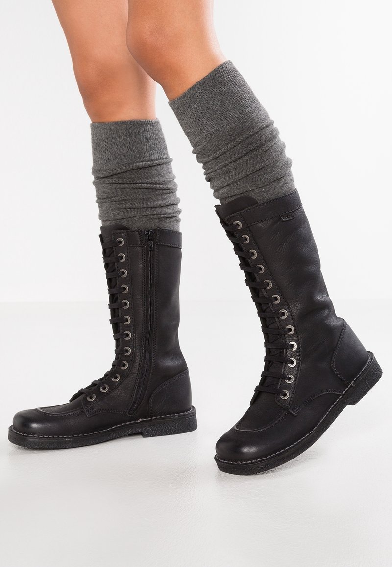 Bottes noires montantes en cuir, avec un laçage à l'avant, une fermeture éclair sur le côté et une semelle en caoutchouc texturé. Portées avec des jambières grises décontractées.