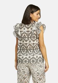 Blusa blanca con intrincados patrones florales negros y mangas con volantes. Combinada con pantalones de estampado que presentan diseños similares.