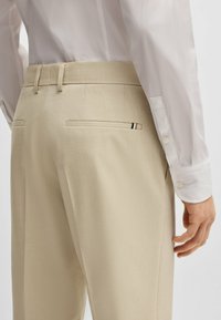 Beige tailored trousers with a smooth texture, featuring two back pockets. A subtle black and white stripe detail is visible on one pocket.