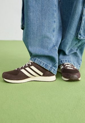 Persona che indossa jeans blu a gamba larga e sneakers marroni con strisce bianche su un pavimento verde, in piedi con i piedi incrociati.