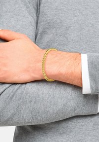 Bracciale in metallo dorato con un design intrecciato, appoggiato su un polso, abbinato a un maglione grigio. Texture liscia e colore uniforme.