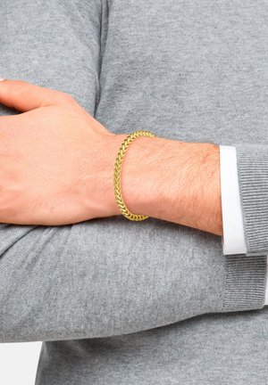 Goldfarbene Metallkette mit geflochtenem Design, die an einem Handgelenk ruht und mit einem grauen Pullover kombiniert ist. Glatte Textur und gleichmäßige Farbe.