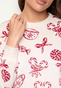 Pitkähihaista puseroa vaaleassa kankaassa, jossa on punaisia makeisteemaisia printtejä, kuten makeispippejä, nauhoja ja juhlakupposia, joissa on lumisateita.