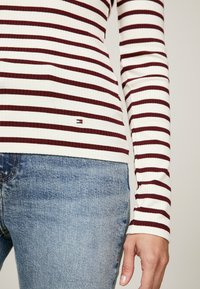 Maglietta a maniche lunghe in tessuto a coste con righe orizzontali bordeaux e bianche, con un piccolo logo del marchio sull'orlo. Jeans di denim visibili sotto.