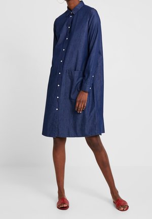 Kvinde iført en løstsiddende mørkeblå langærmet knap-op kjole med forlommer og røde slip-on sandaler, stående med det ene ben fremad.