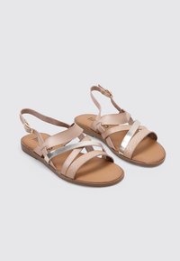 Sandalias beige con múltiples correas entrecruzadas, que incluyen acentos plateados brillantes y un diseño texturizado en la plantilla. Suela plana.