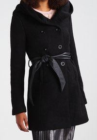 Cappotto nero doppiopetto con cappuccio, dotato di una cintura in materiale lucido e tessuto testurizzato, presentato con due tasche frontali.
